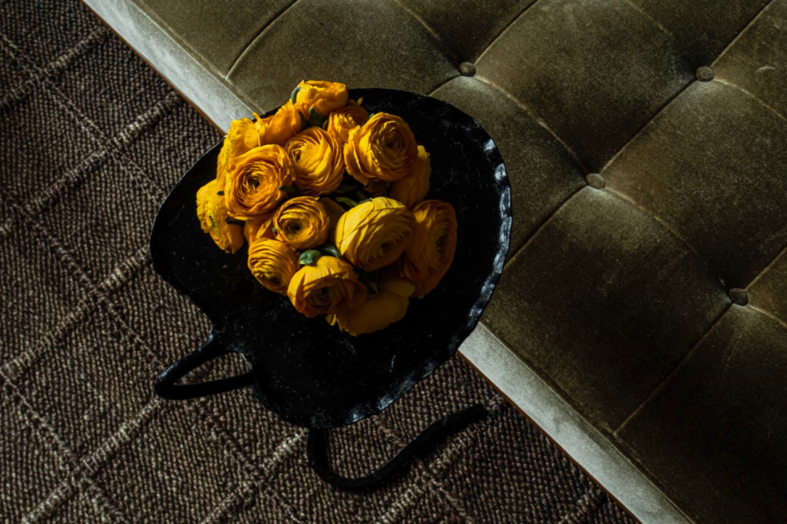 Yellow ranunculus on a black iron side table beside an olive velvet bench